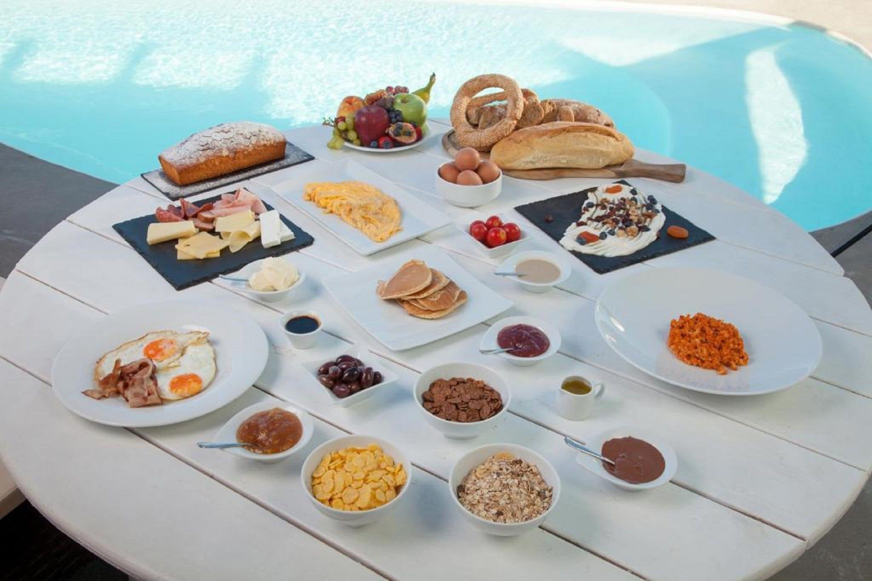 Un buffet de petit-déjeuner est dressé sur une table blanche, près d’une piscine, et propose une variété de plats et de boissons.