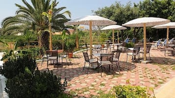 Uma área de pátio ao ar livre com piso de azulejos hexagonais, várias mesas e cadeiras, e guarda-sóis de palha oferecendo sombra.
