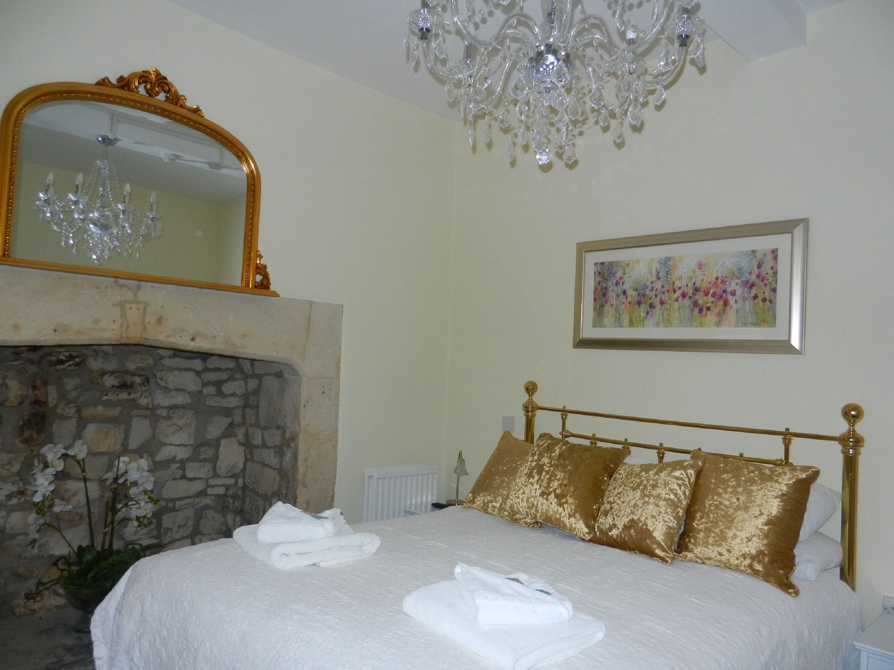 Un dormitorio con chimenea de piedra, un candelabro, una cama con cabecera y almohadas doradas, un cuadro enmarcado y un espejo con marco dorado.