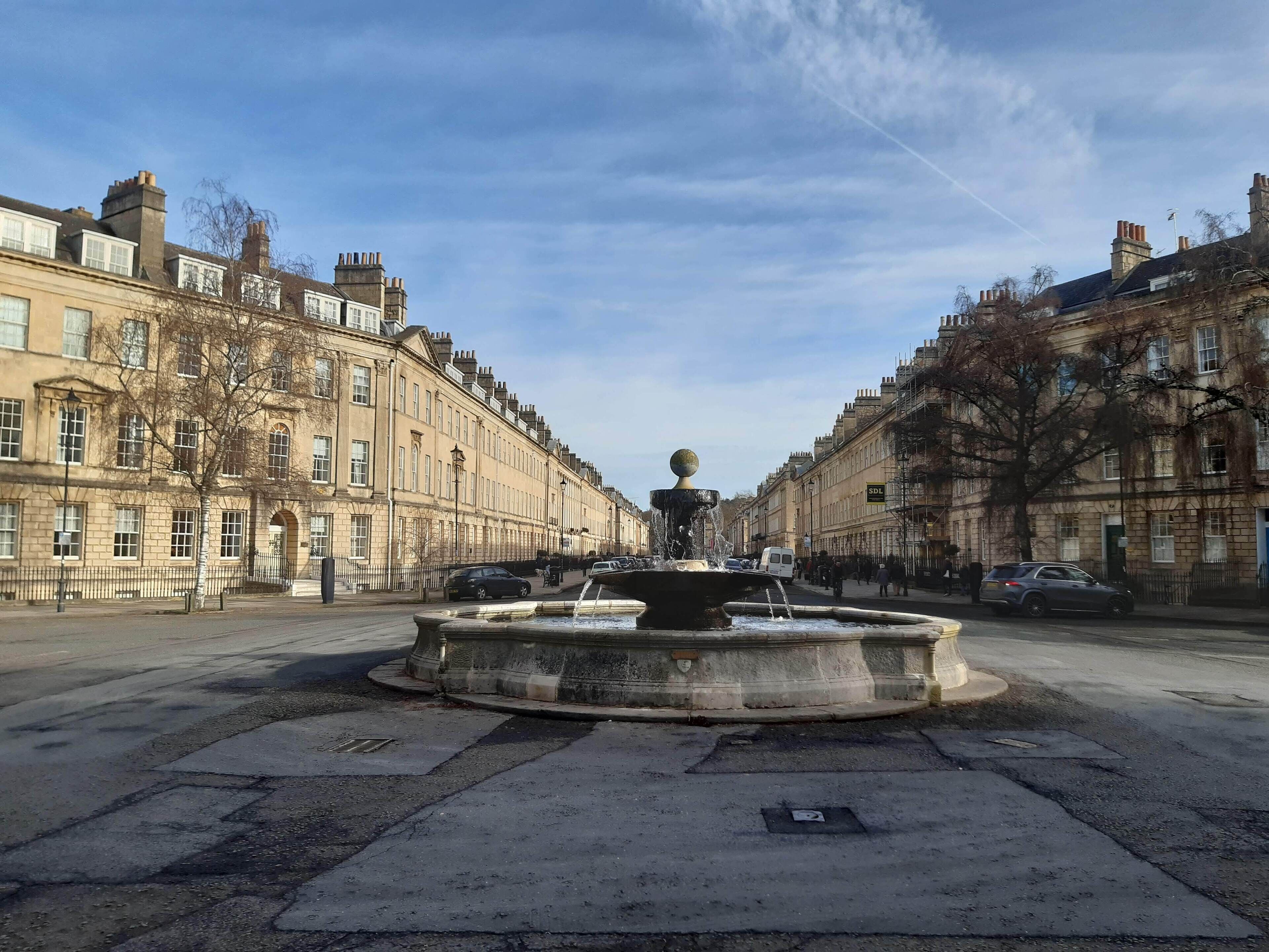 Una calle histórica con una fuente central, flanqueada por edificios tradicionales y un cielo despejado.