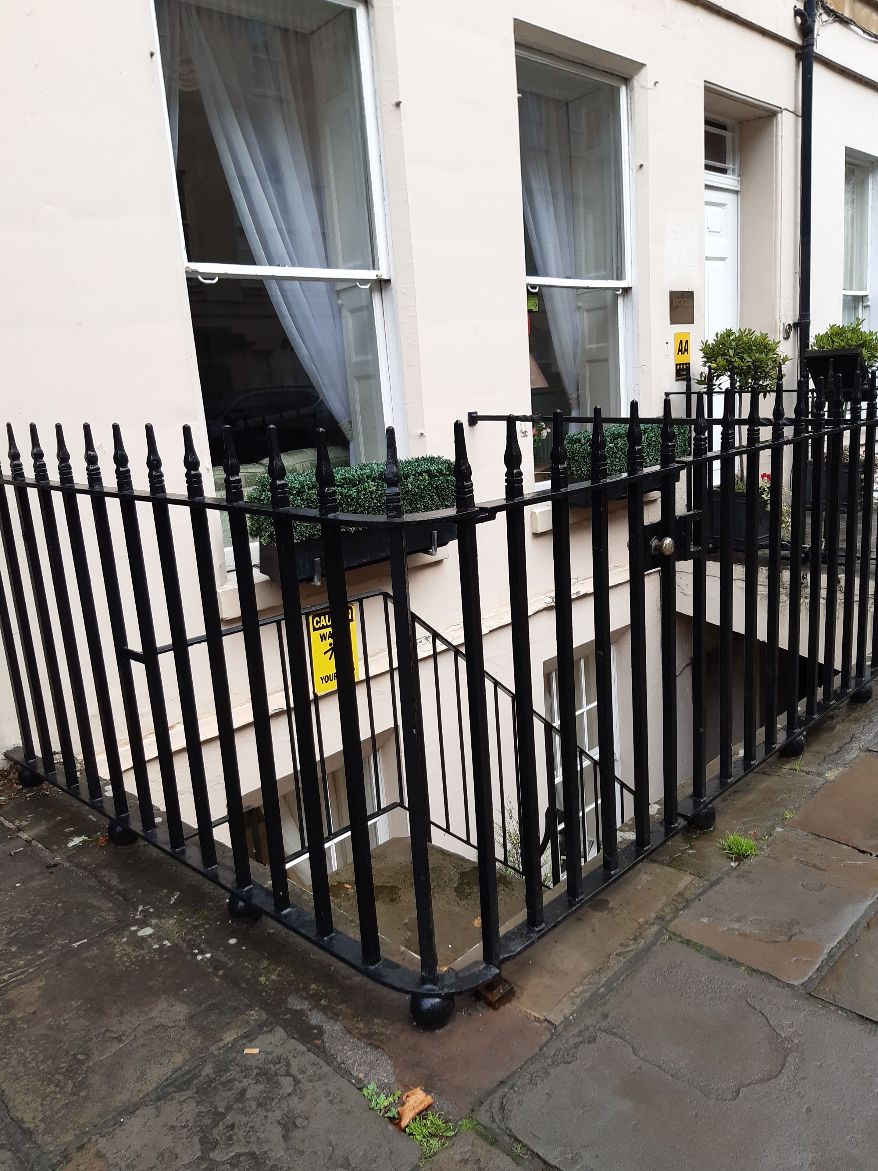 A residential building with a black metal fence, windows with white curtains, and a yellow caution sign.