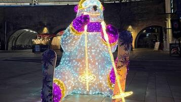 Una persona vestida con un disfraz iluminado, con un elemento central brillante, de pie en un entorno urbano con un túnel y edificios al fondo.