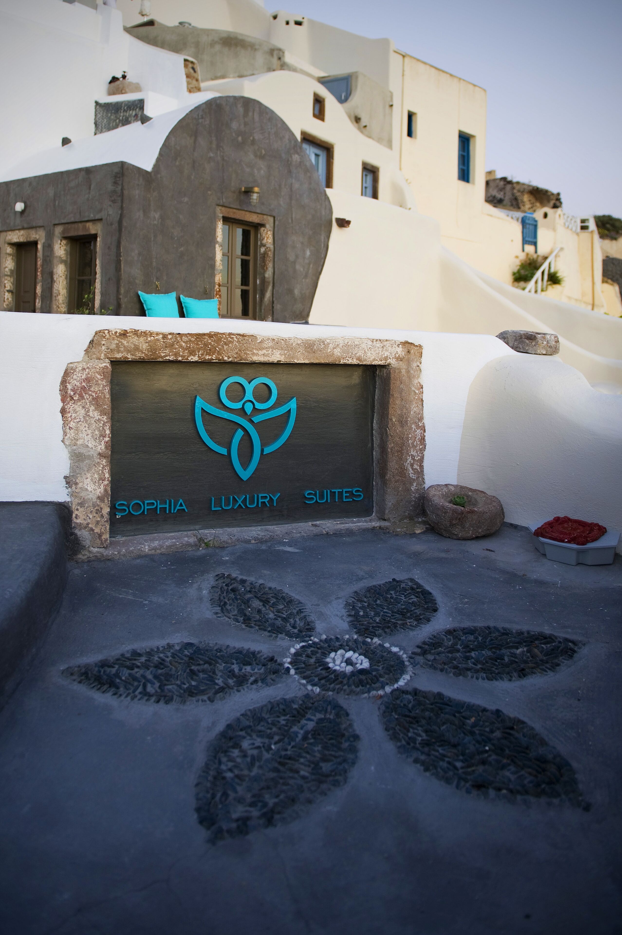 Une enseigne pour le Sophia Luxury Suites, avec un logo, un chemin de pierre menant à l’entrée, et un bâtiment à l’architecture blanche et grise en arrière-plan.