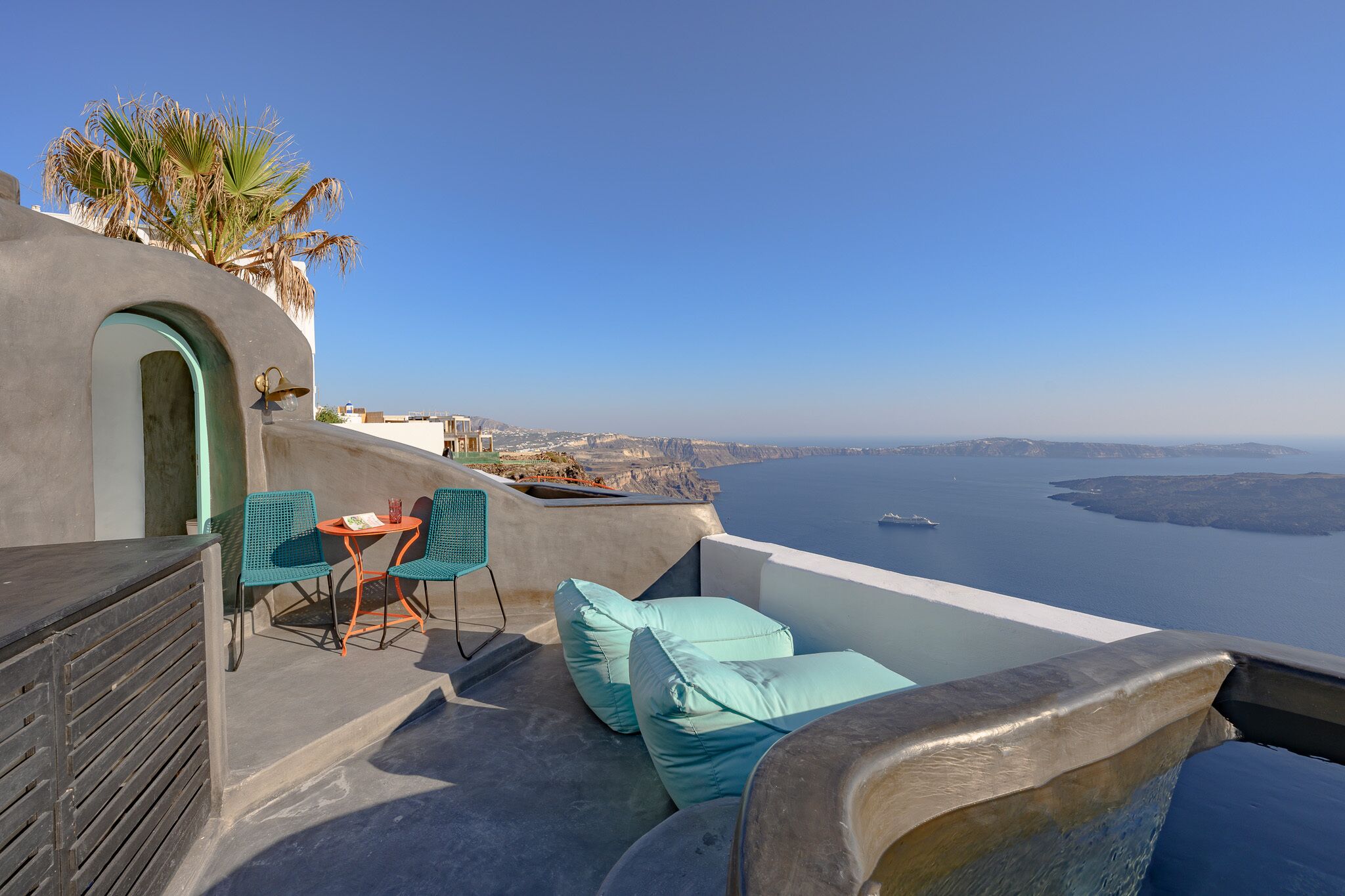 A terrace with a view of the sea, featuring a table and chairs, a small seating area with cushions, and a palm tree.