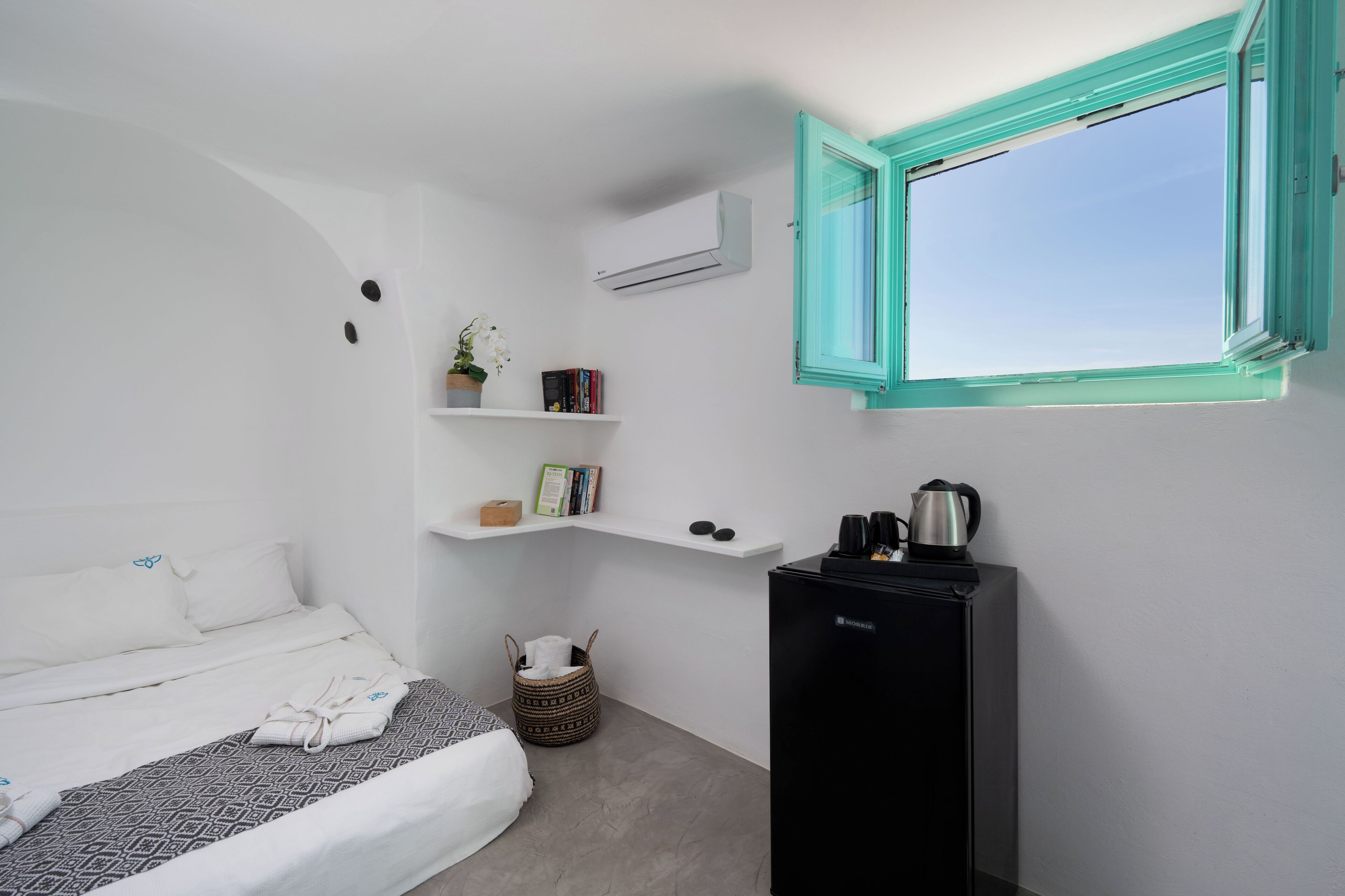 Un dormitorio pequeño con una cama, un estante bajo con libros y una planta, una ventana con marcos verde azulado, y un refrigerador negro.