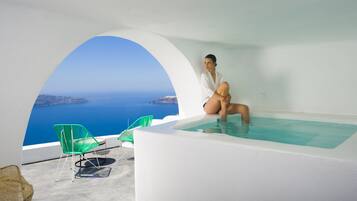 A person sitting in a white infinity pool with a view of the sea and islands. There are green chairs and a white wall.