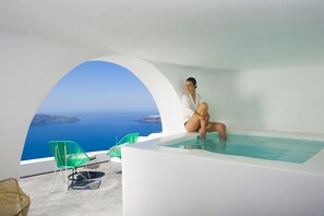 A person sitting in a white infinity pool with a view of the sea and islands. There are green chairs and a white wall. - Sophia Luxury Suites (Santorini)