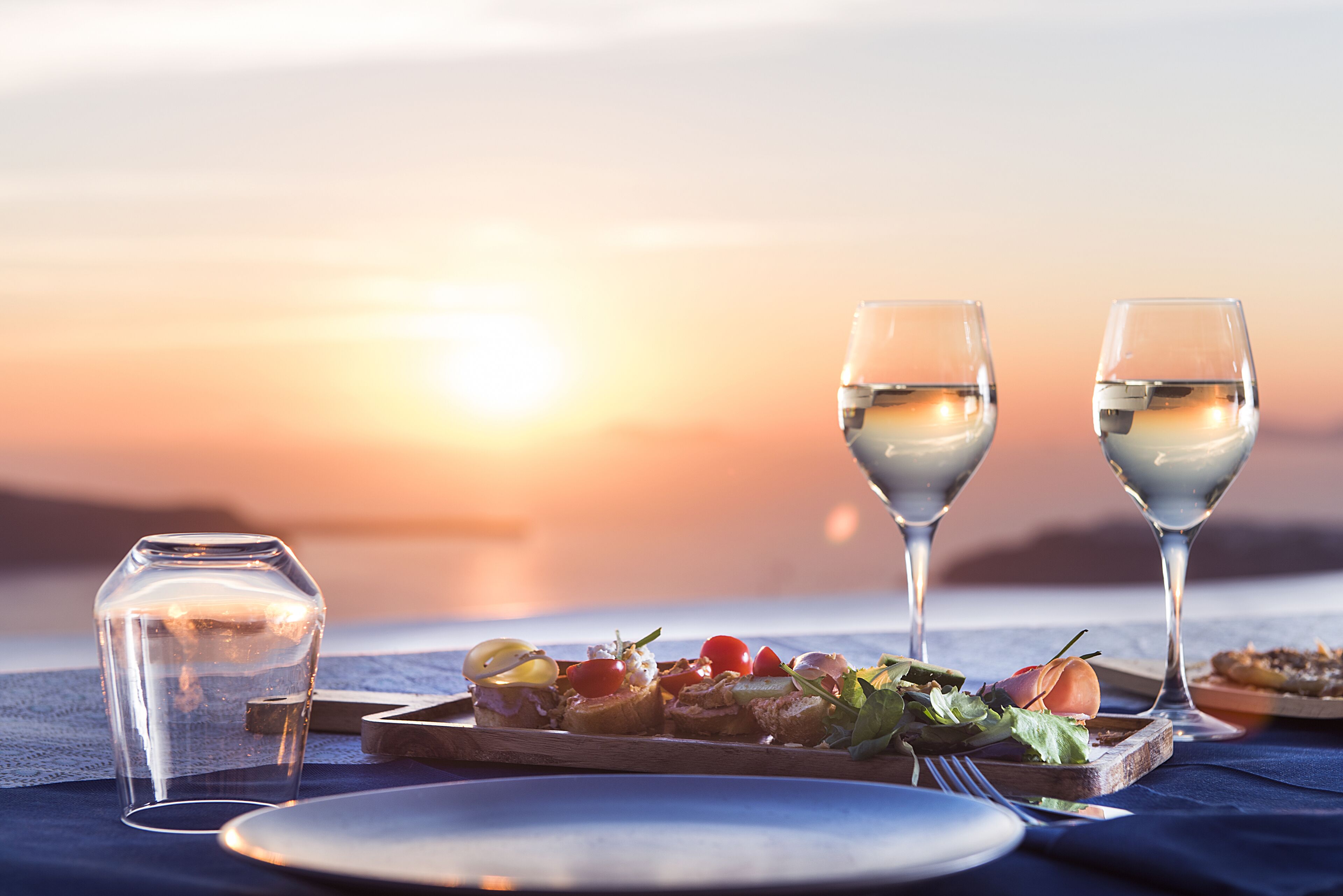 Une table dressée pour un repas, avec une vue sur le coucher de soleil, deux verres à vin et un verre d’eau.
