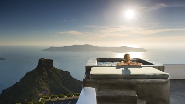 A person relaxing in a sunlit outdoor bathtub with a view of the sea and distant islands.