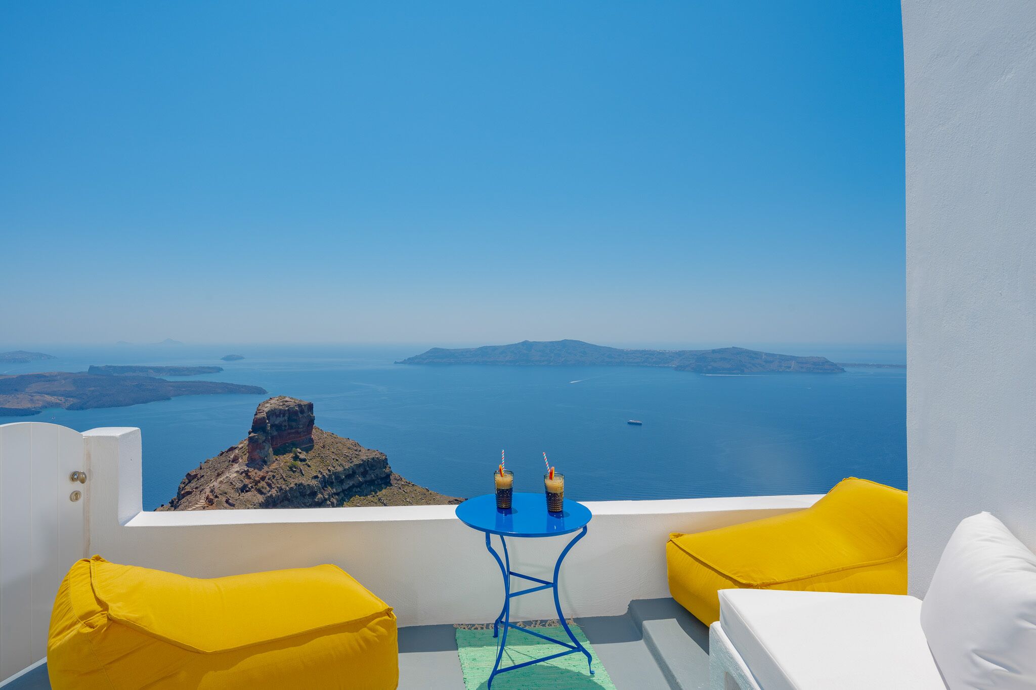 A balcony with a white wall, a blue table with two bottles, yellow cushions, and a view of the sea and islands.