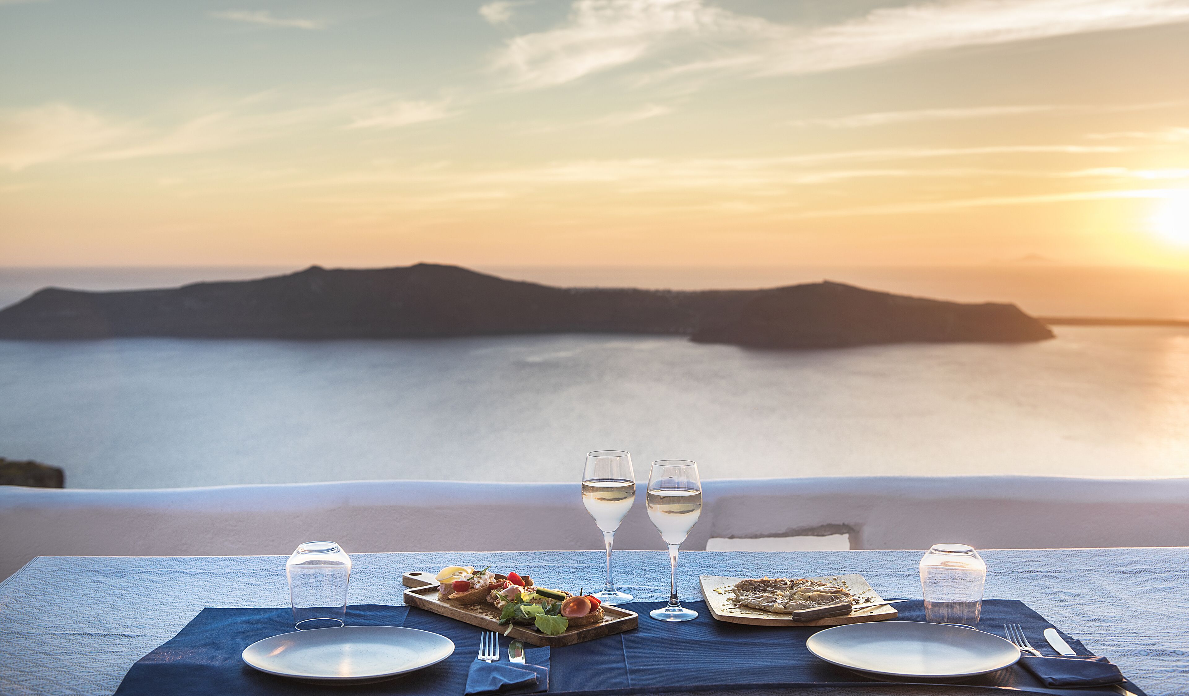 A table set with plates, cutlery, and glasses, overlooking a coastal landscape with mountains and the sea at sunset.
