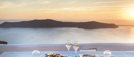 A table set with plates, cutlery, and glasses, overlooking a coastal landscape with mountains and the sea at sunset.