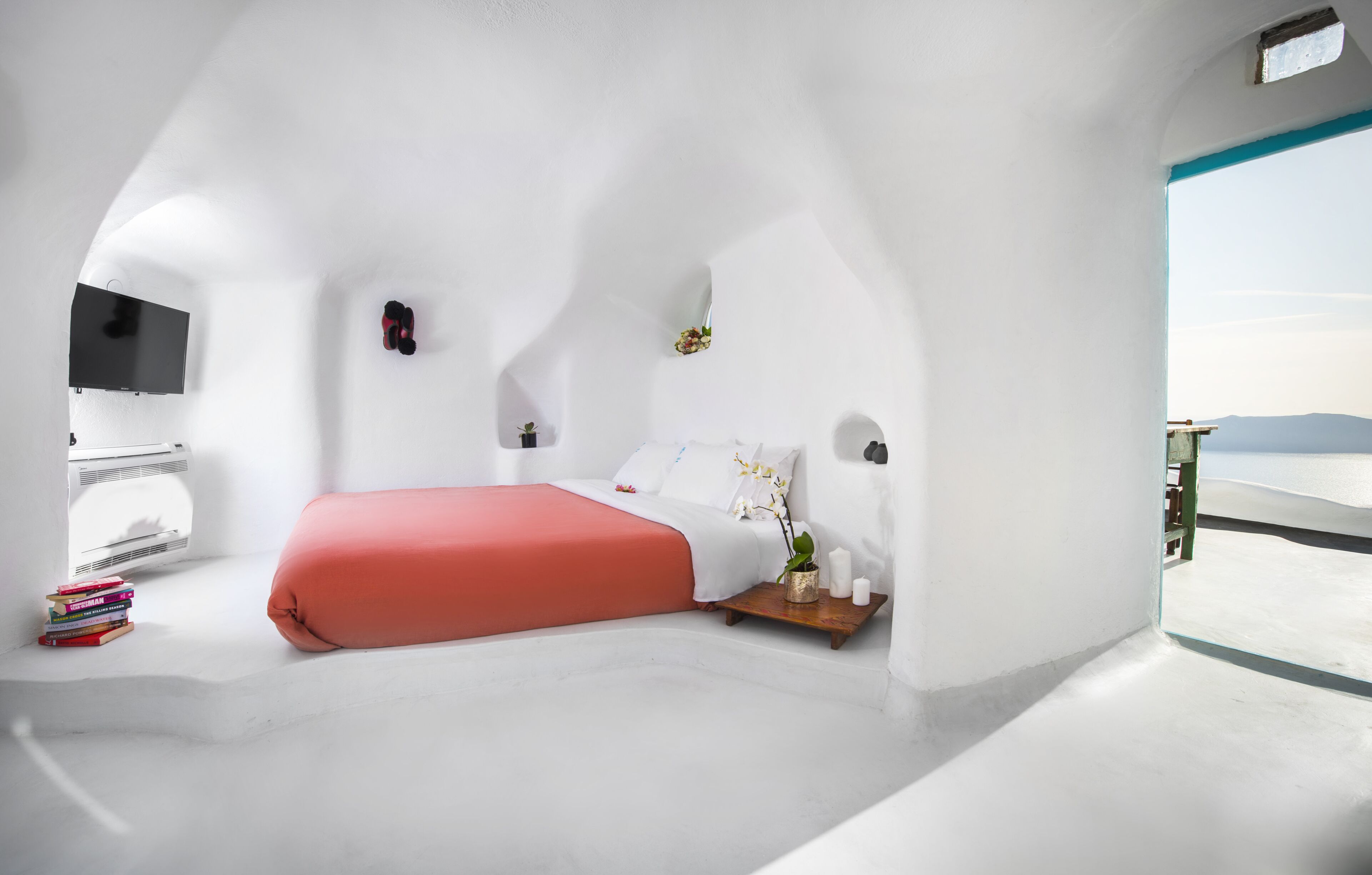 Una habitación blanca y curva con una cama, un televisor y una mesita con libros y una planta.