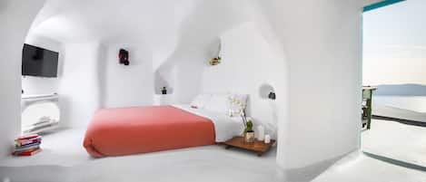 Una habitación blanca y curva con una cama, un televisor y una mesita con libros y una planta.
