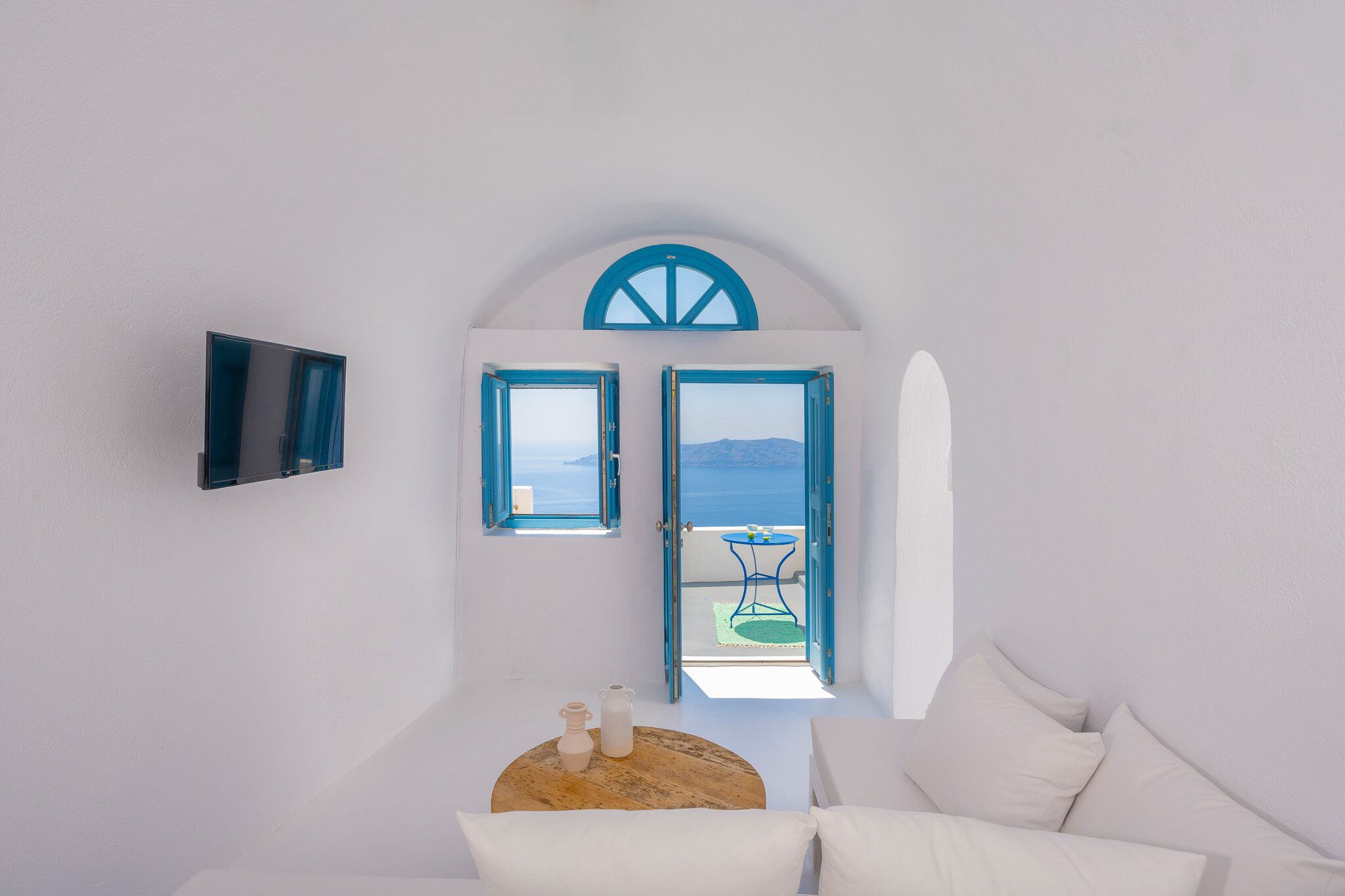 Um quarto com parede branca, uma mesa de madeira com duas garrafas brancas, um sofá branco e uma TV fixada na parede.