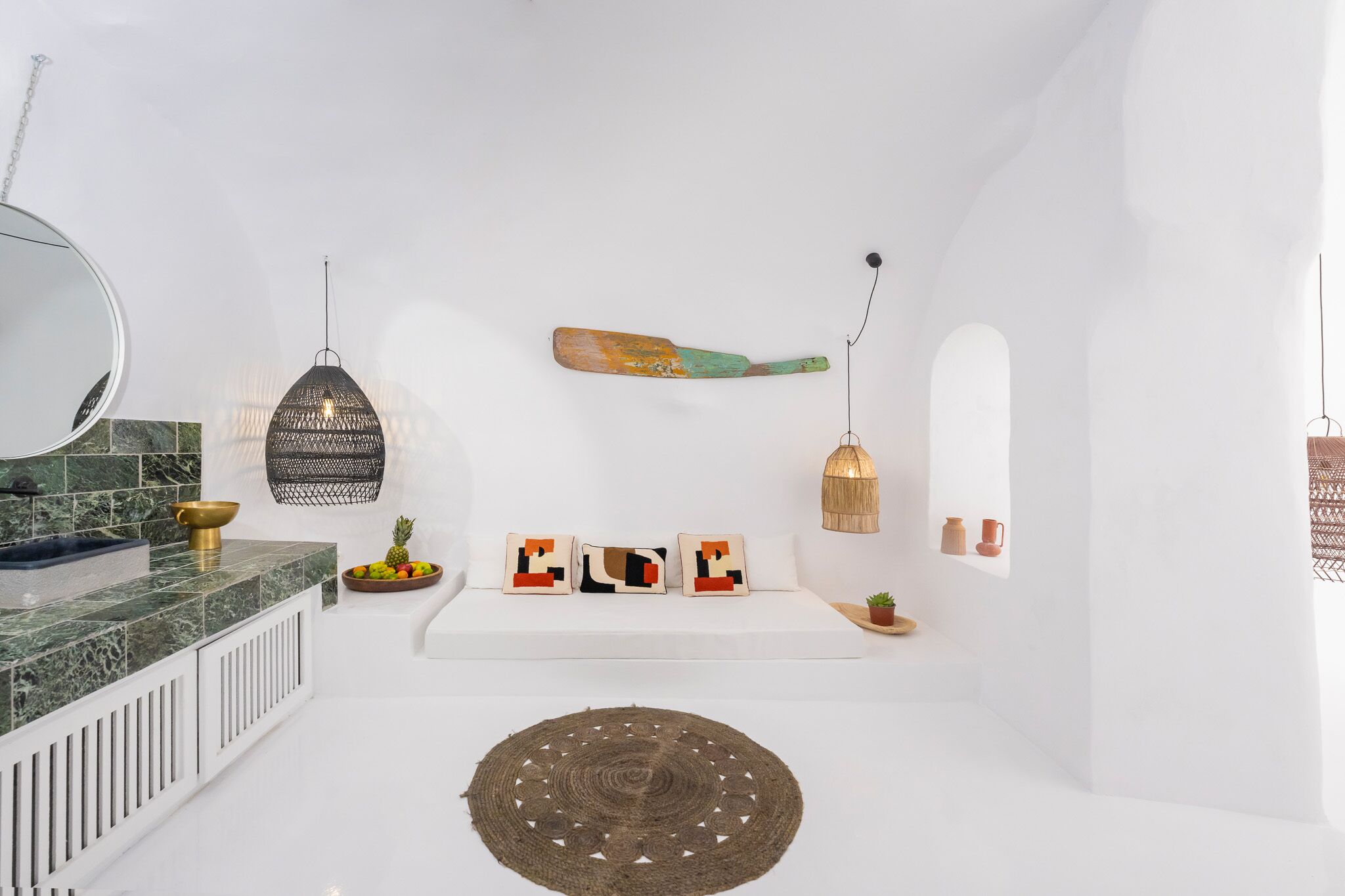 A modern bathroom with a white sink, a round mirror, a hanging light fixture, a decorative wall hanging, and a small plant.