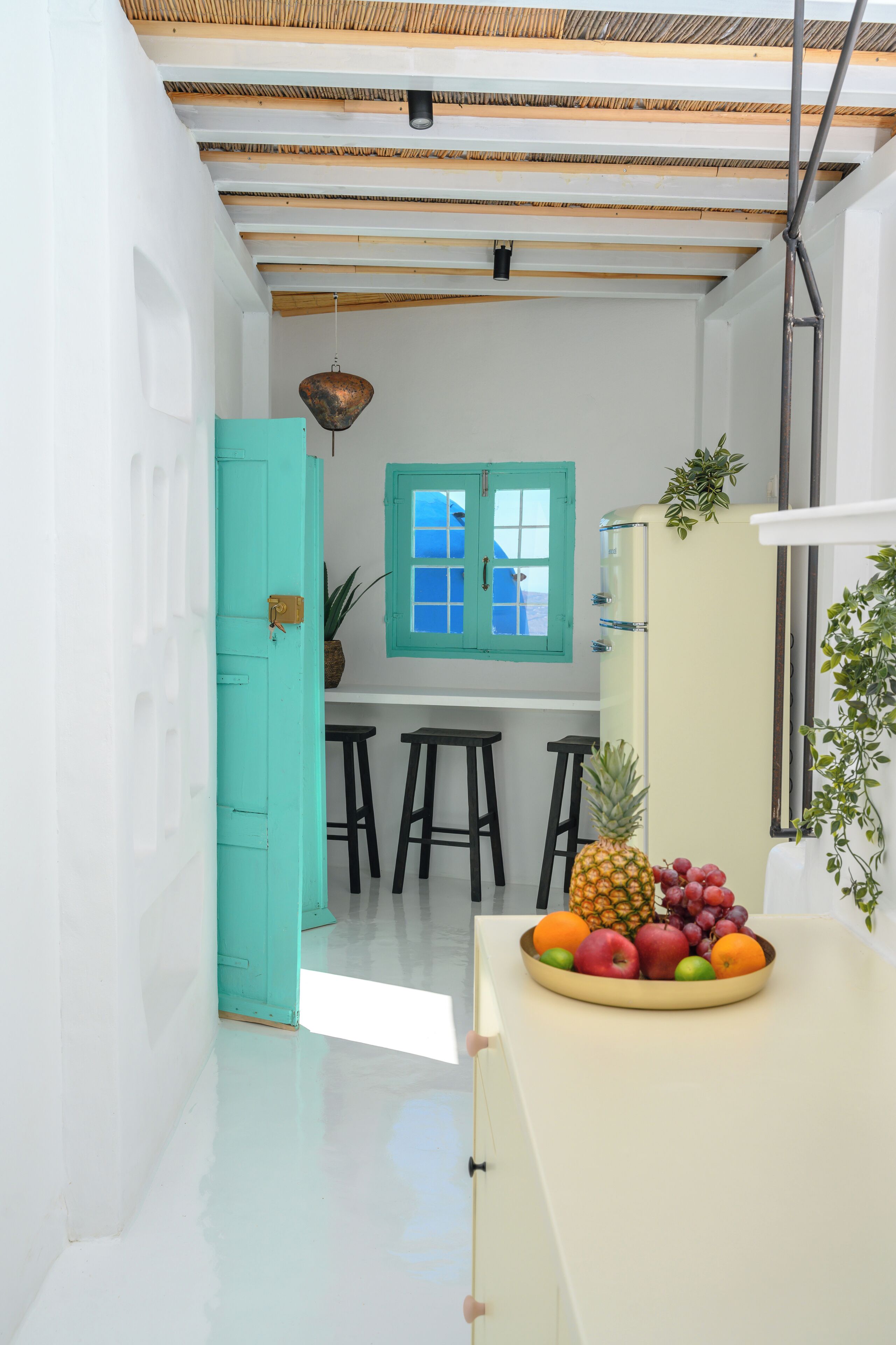 A narrow kitchen with a teal door, a window with blue shutters, and a fruit bowl on the counter.