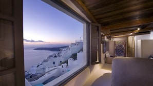 A room with a large window overlooking a coastal village, wooden ceiling, and a bed with a white headboard.