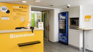 A hotel reception area with a yellow wall displaying room rates and amenities. There is a reception desk with a 'Bienvenue' sign, a blue kiosk with a 'KLIK' sign, and a microwave in the background. - hotelF1 Saint Etienne La Grand Croix (La Grand-Croix)
