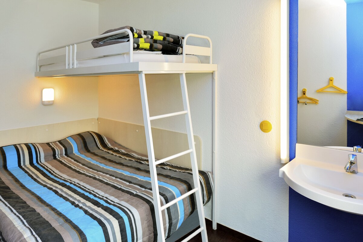Een stapelbed met een ladder, een nachtkastje, een wandlamp, en een badkamer met een wastafel en spiegel.