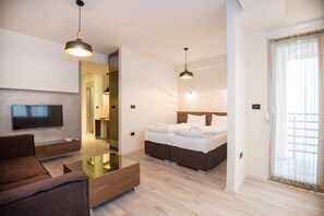 A modern hotel room with a flat-screen TV, a sofa, a glass coffee table, a bed with white linens, and pendant lighting.