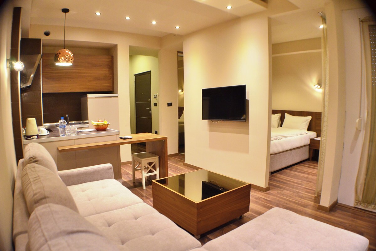 A modern hotel room with a sofa, a glass coffee table, a TV, a bed, and a small kitchenette.