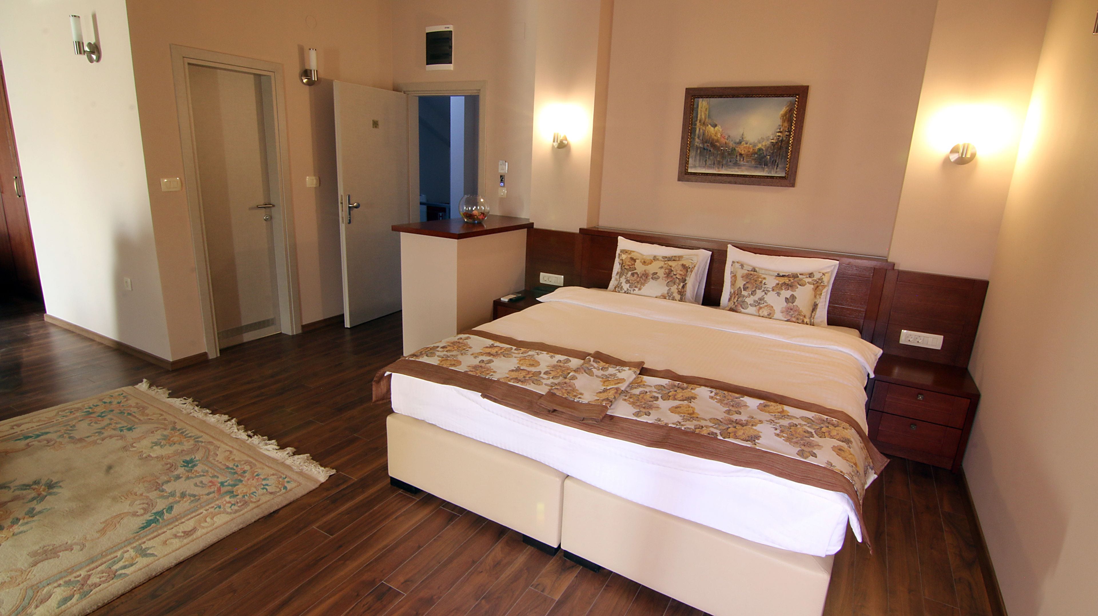 A bedroom with a large bed, bedside tables, a nightstand, a painting on the wall, and a door leading to another room.