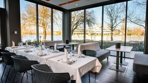 Un restaurante con mesas dispuestas para comer, grandes ventanales que ofrecen vistas a un cuerpo de agua y edificios.