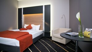 Una habitación de hotel con una cama, una silla, una mesa y un florero con una flor.