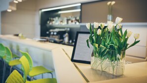 Un área de recepción con un jarrón de vidrio transparente que contiene flores blancas sobre un mostrador, un atril para menús y sillas verdes.