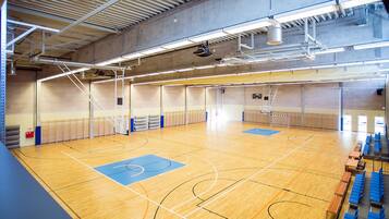 An indoor basketball court with a wooden floor, blue and black markings, and seating along the sides.