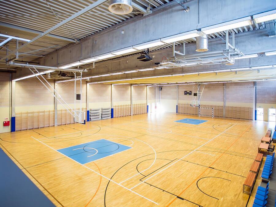 An indoor basketball court with a wooden floor, blue and black markings, and seating along the sides.