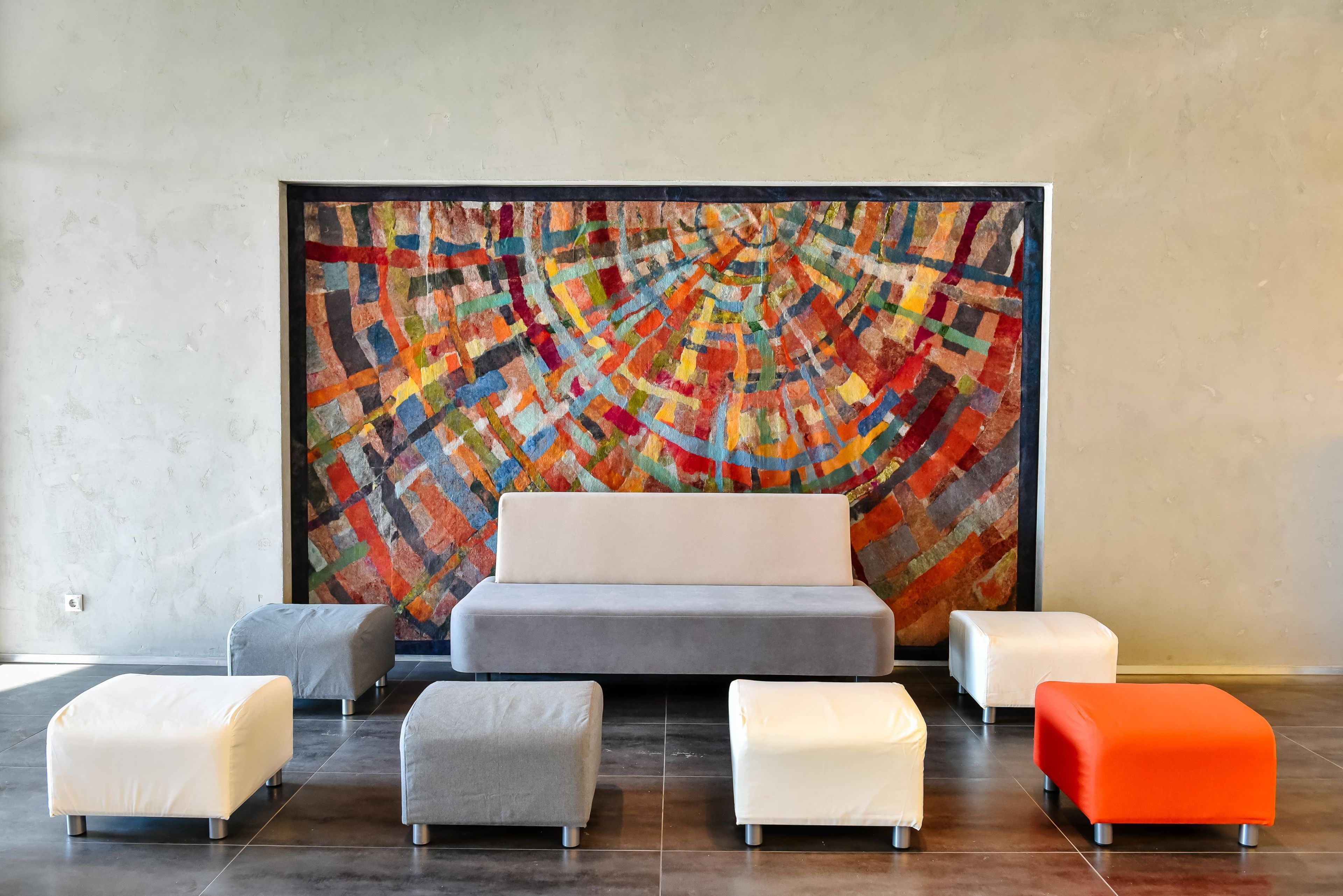 A modern room with a large abstract painting on the wall, a grey sofa, and four ottomans in white and orange.