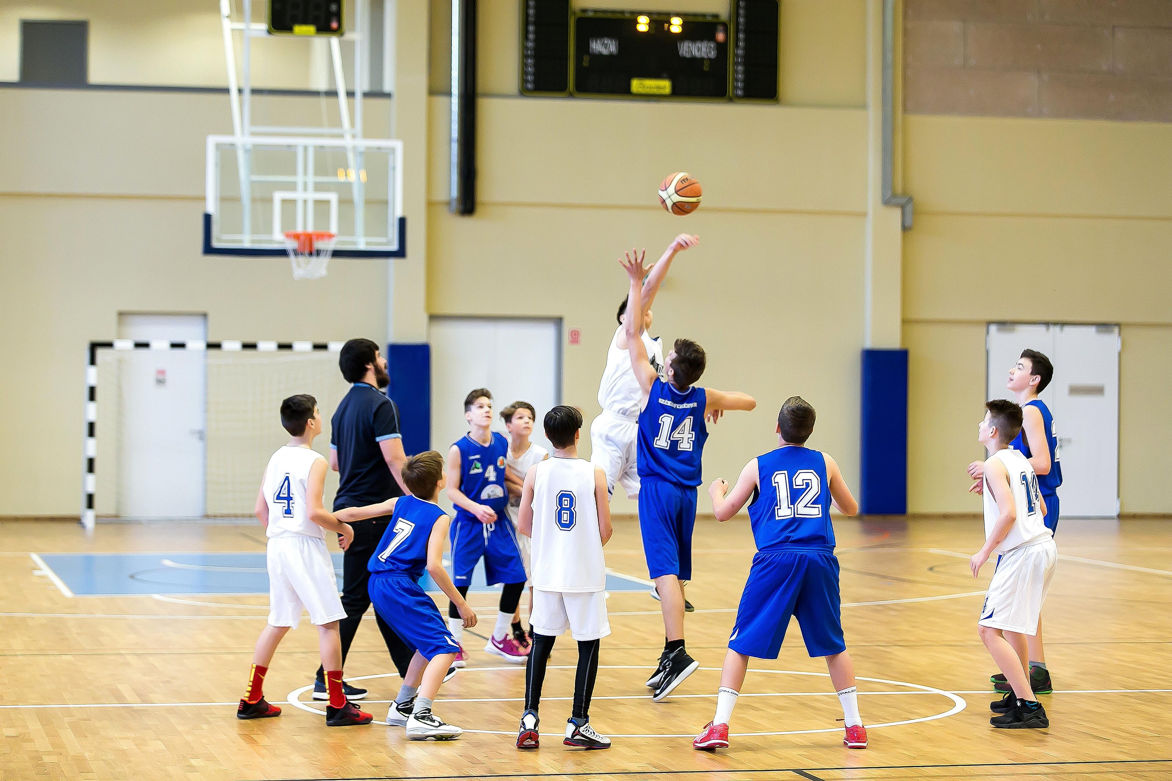 A basketball game in a gymnasium with a basketball hoop, players in blue and white uniforms, and a scoreboard in the background.