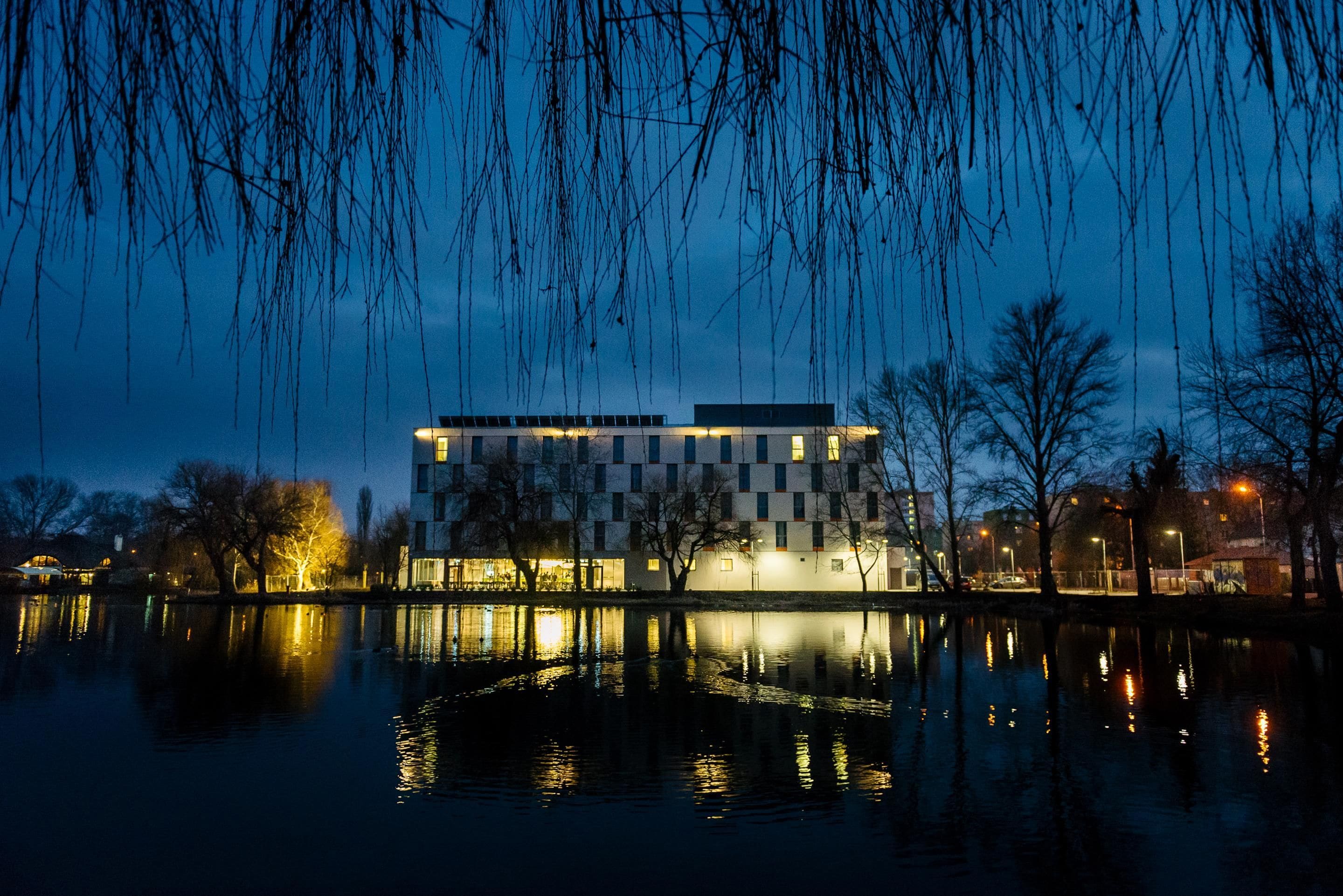 A modern building with a checkered facade, reflecting on a calm body of water, surrounded by bare trees and a clear sky.