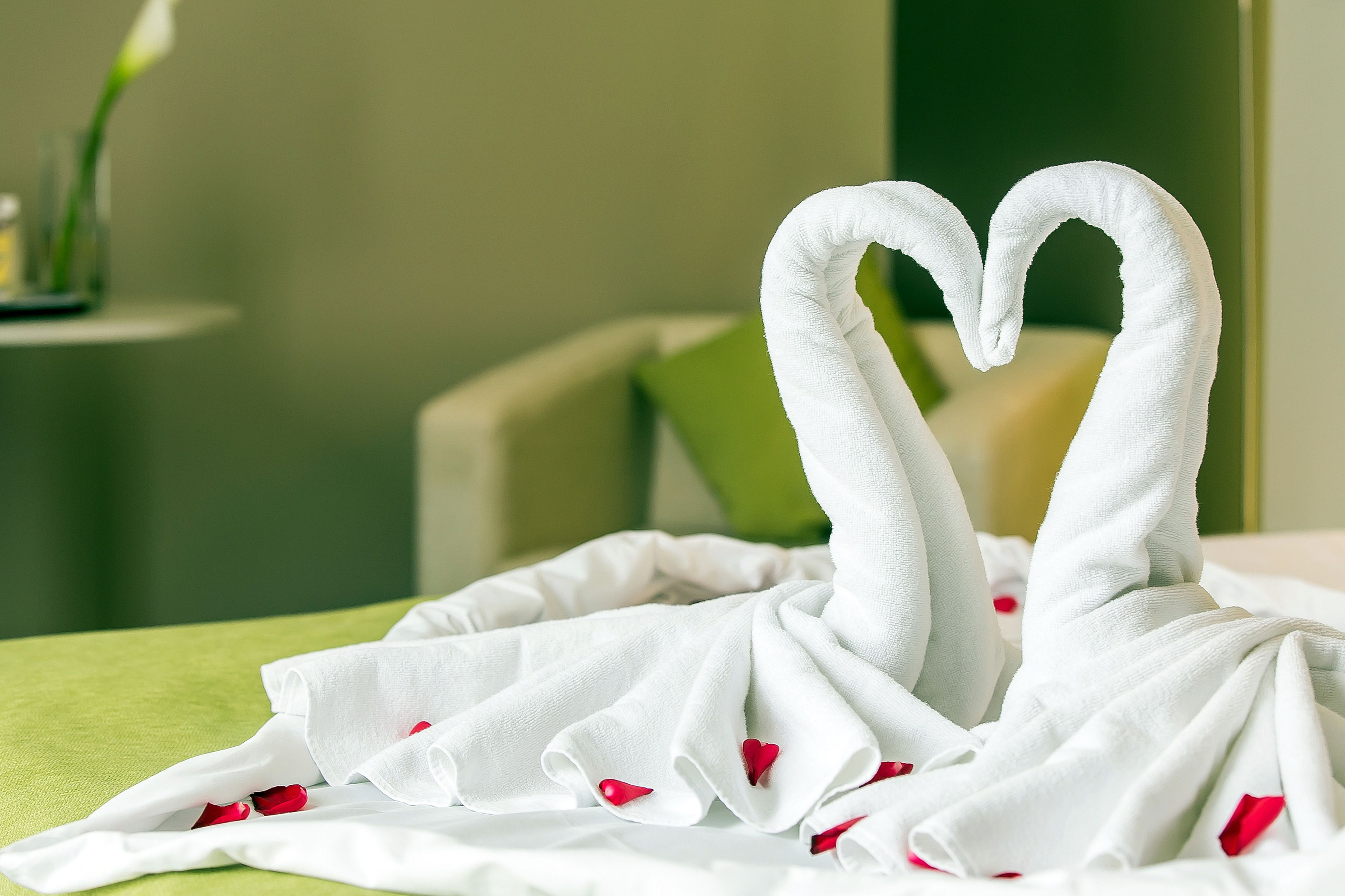 Ein Bett mit weißen Laken und roten Rosenblättern, eine Vase mit einer Blume und eine Handtuch-Arrangement in Herzform.
