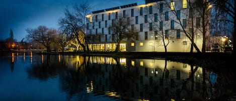Un edificio moderno con una superficie acuática reflectante en primer plano, que presenta una combinación de ventanas rectangulares y cuadradas.