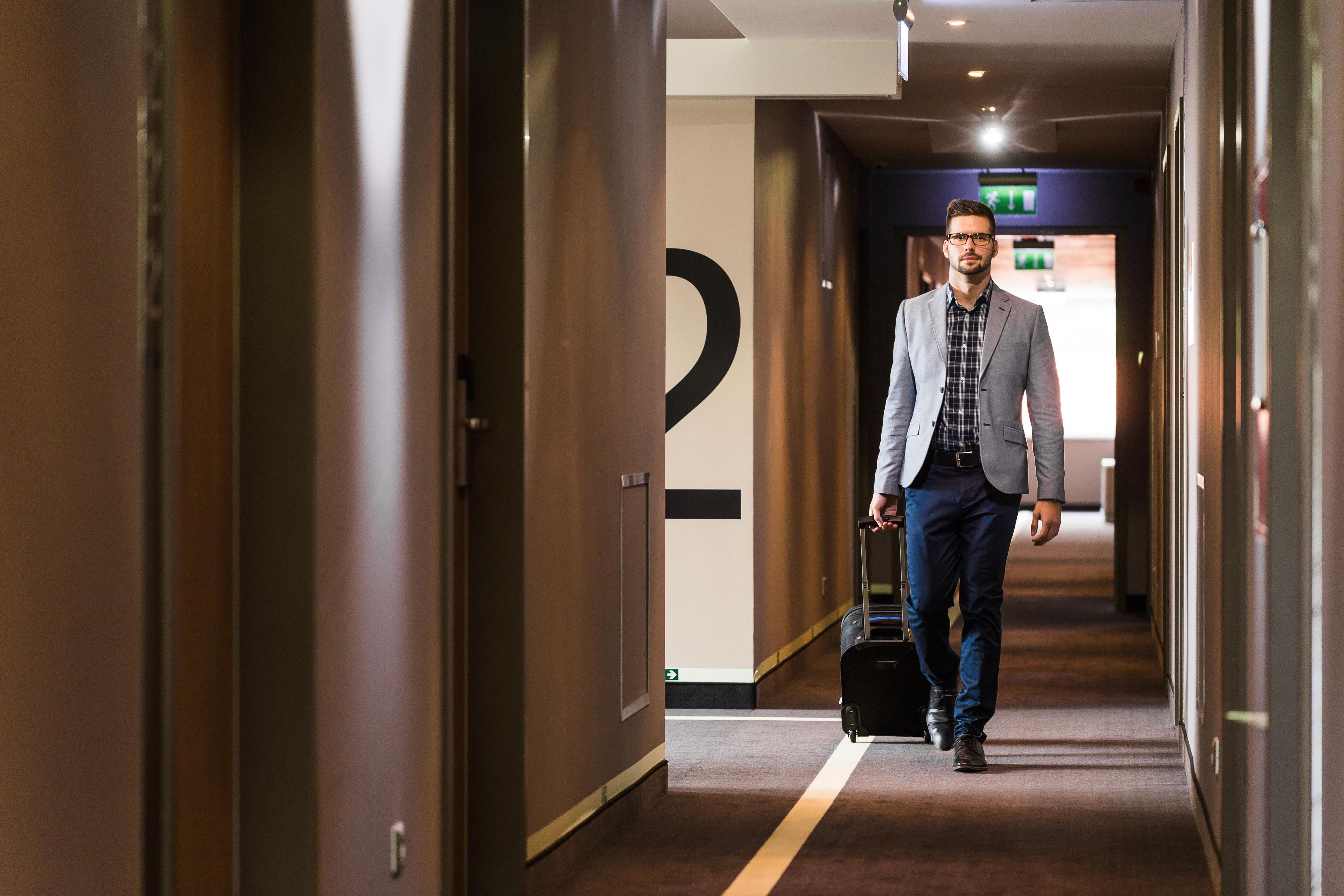 A man walking down a hotel corridor with a suitcase, a sign with a number, and a door on the right.