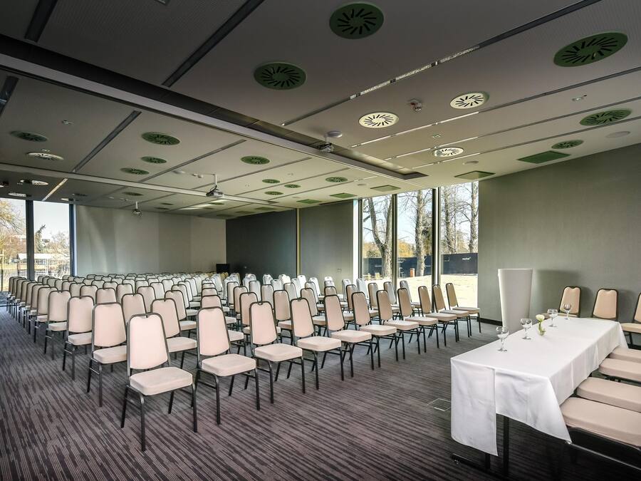 Una sala de conferències amb files de cadires, una taula llarga amb estovall i grans finestres que donen vistes a arbres.