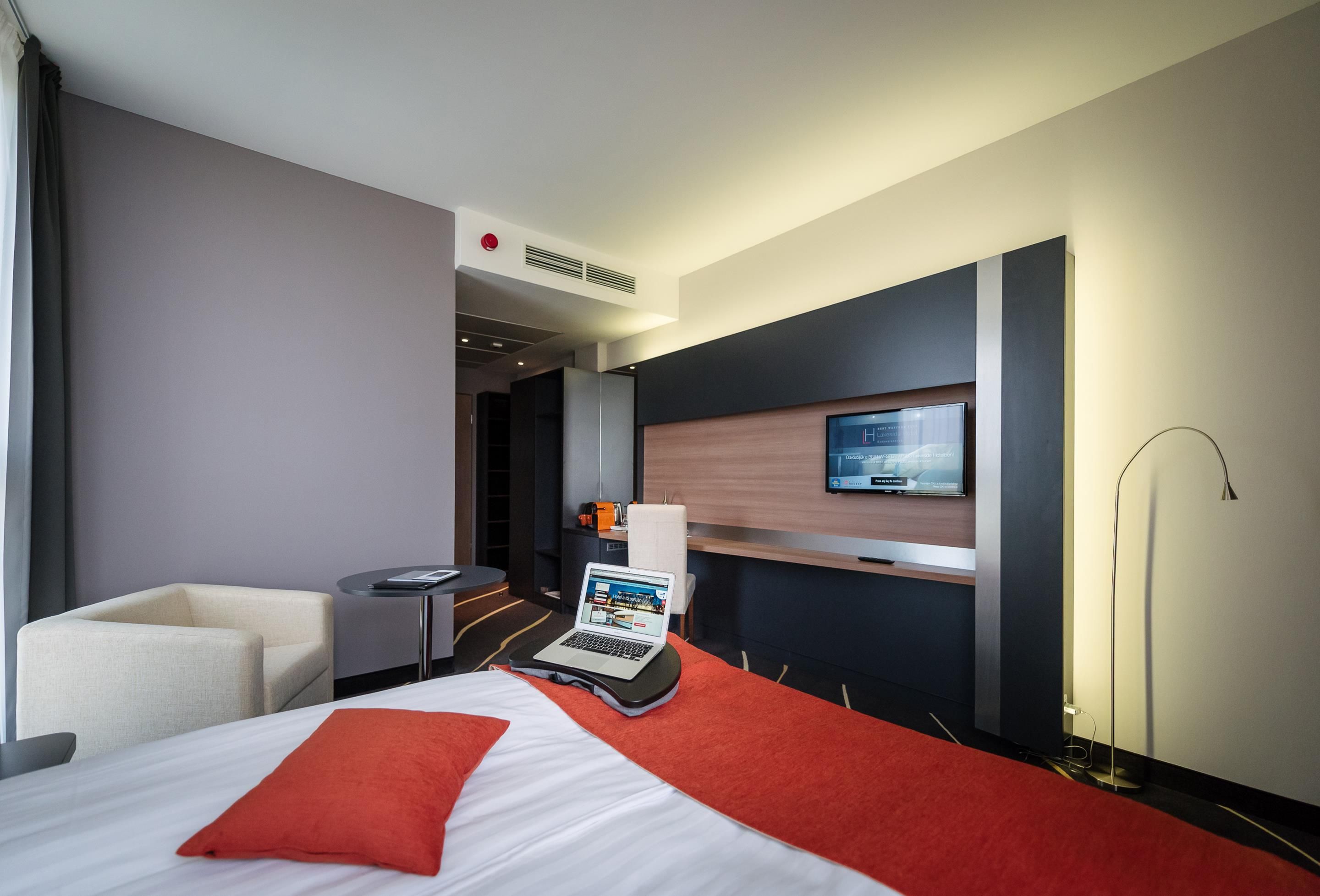 A modern hotel room with a bed, a desk with a laptop, a TV mounted on the wall, and a chair.