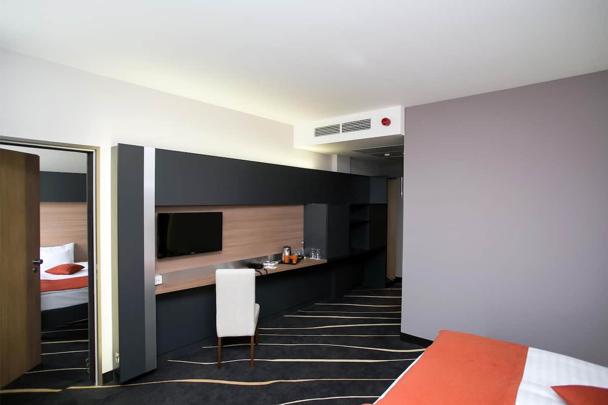 A hotel room with a bed, a desk with a flat-screen TV, a chair, and a small table with a kettle and cups.