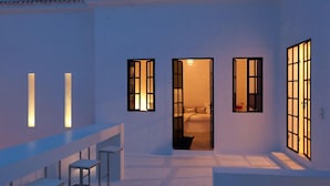 Un intérieur moderne avec un long banc et deux tables, une porte donnant sur une chambre à coucher, et trois fenêtres éclairées.