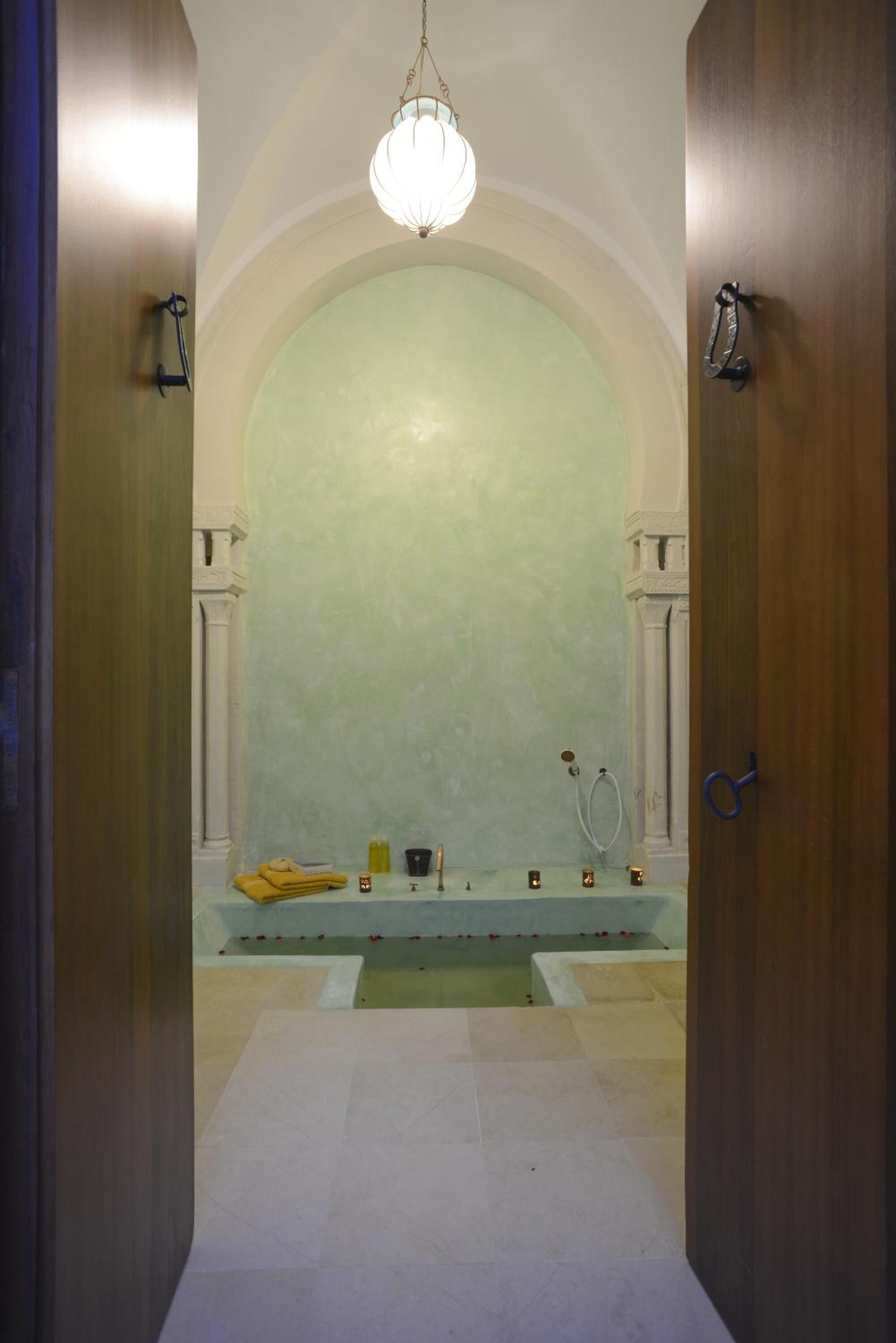 A hallway leading to a room with a large arched doorway, a chandelier, and a bench with a yellow towel and a cup.