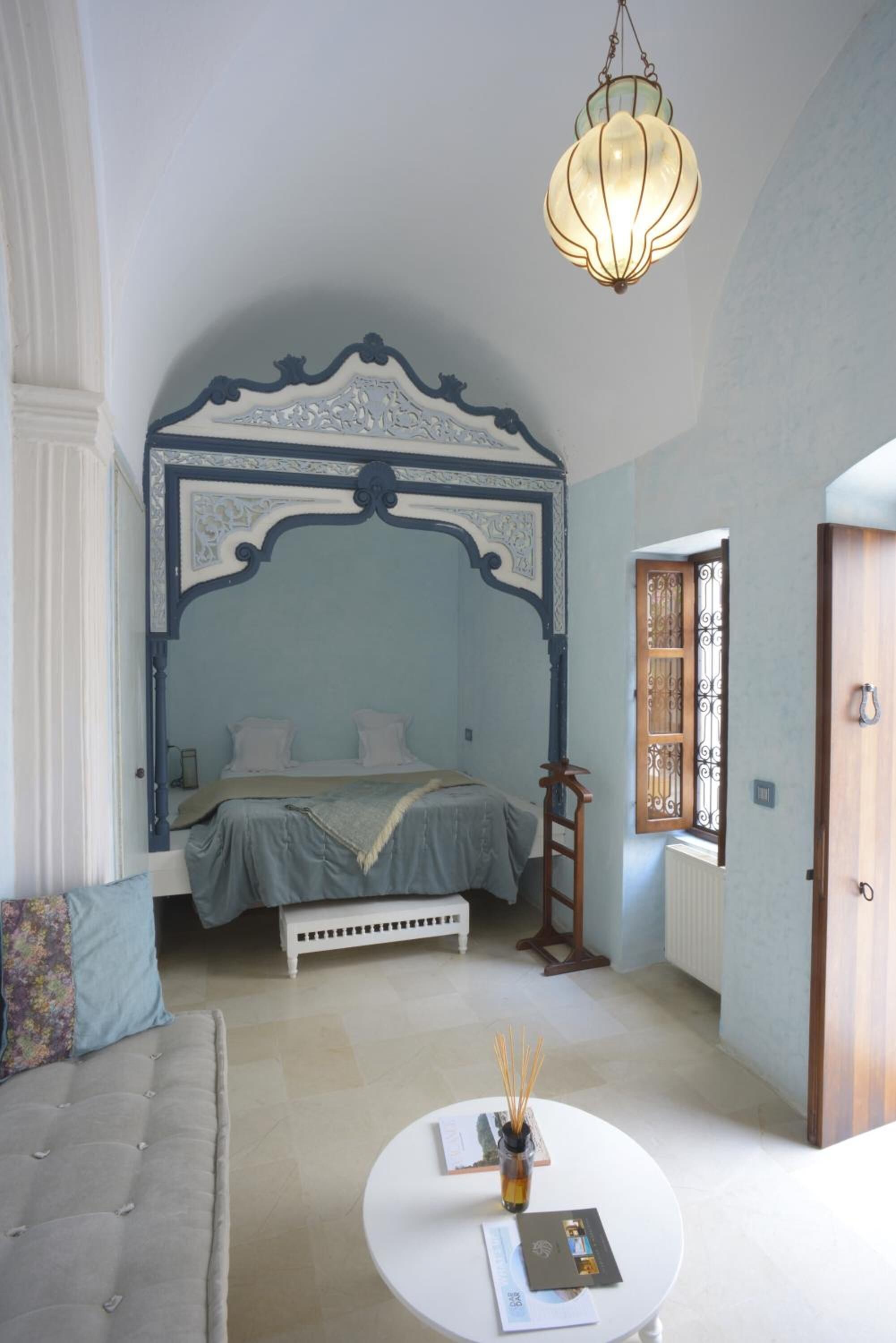 Un dormitorio con una cama con dosel, una mesa redonda con un florero y una ventana con celosía decorativa.
