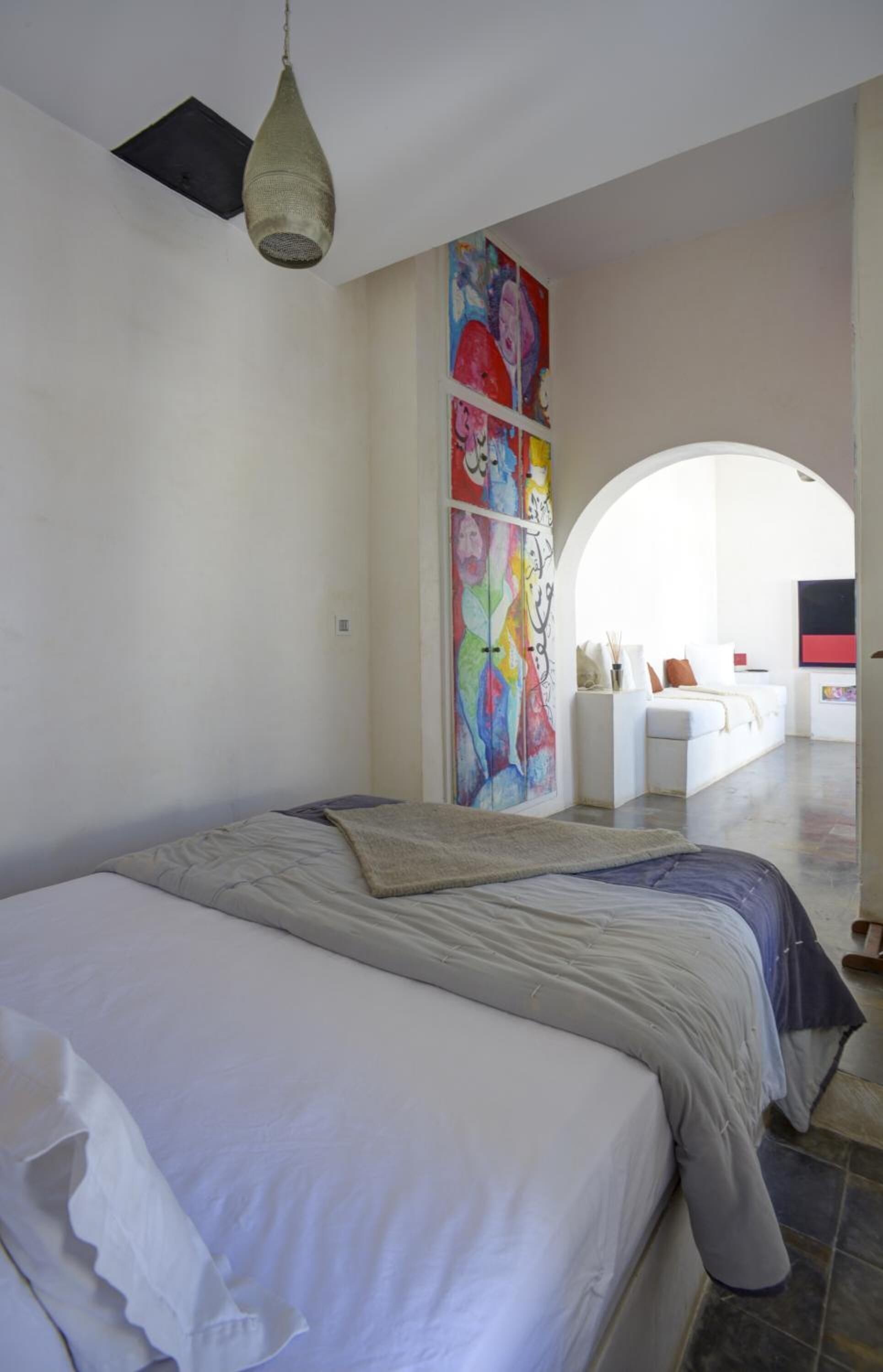 A bedroom with a bed, a ceiling fan, a colorful abstract painting, and a white sofa in the background.