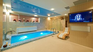 A modern indoor swimming pool with a glass-enclosed shower area, a comfortable lounge chair, and a mounted TV screen. - Boutique Hotel Buta (Minsk)