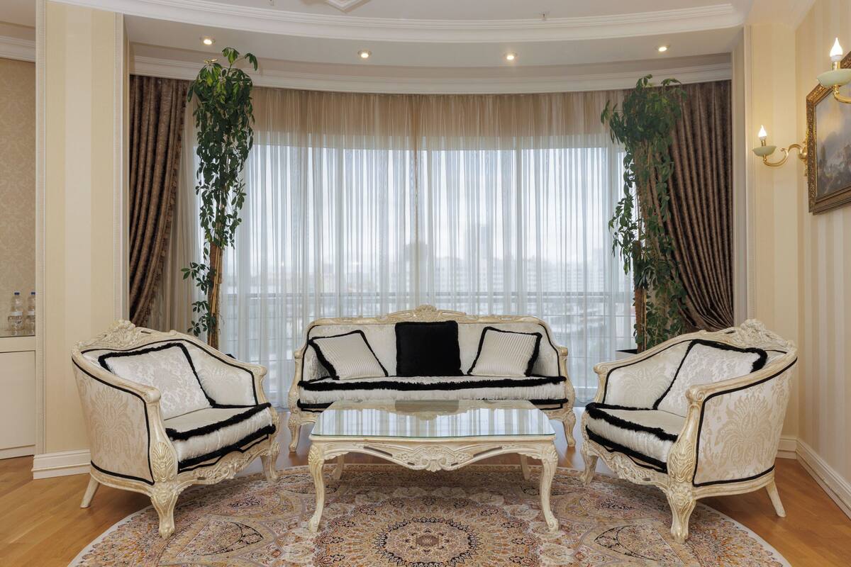 A room with a white sofa set, two armchairs, a patterned rug, and a large window with curtains.