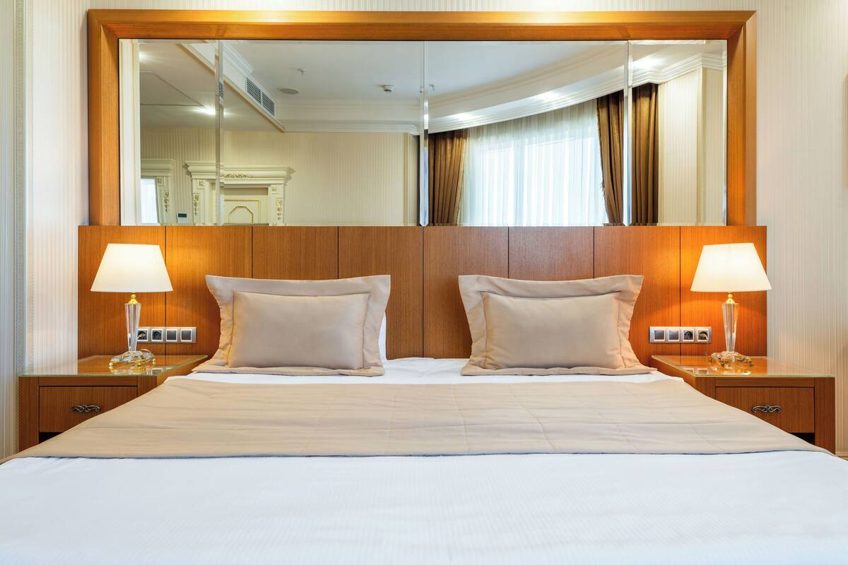 A hotel room with a large bed, two bedside tables with lamps, a large mirror above the bed, and a wooden headboard.