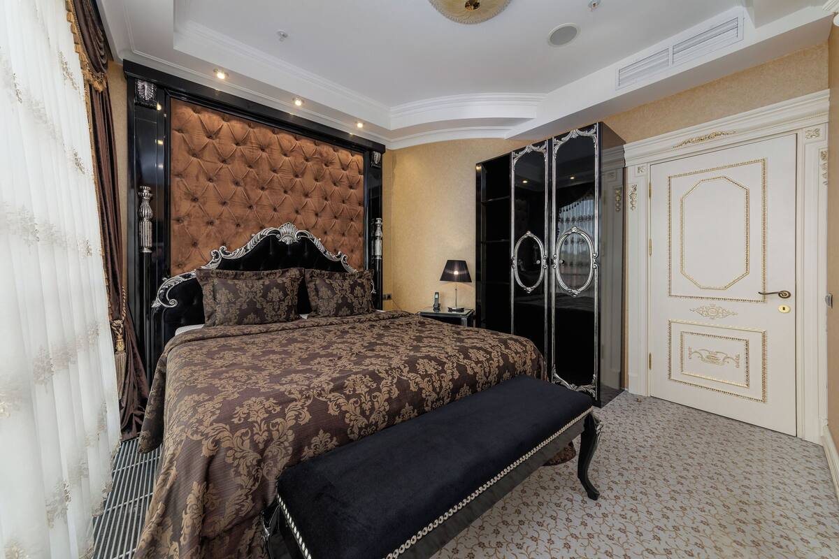 A bedroom with a tufted headboard, a large bed with a patterned bedspread, a bench, a wardrobe with a mirror, and a door with decorative gold detailing.