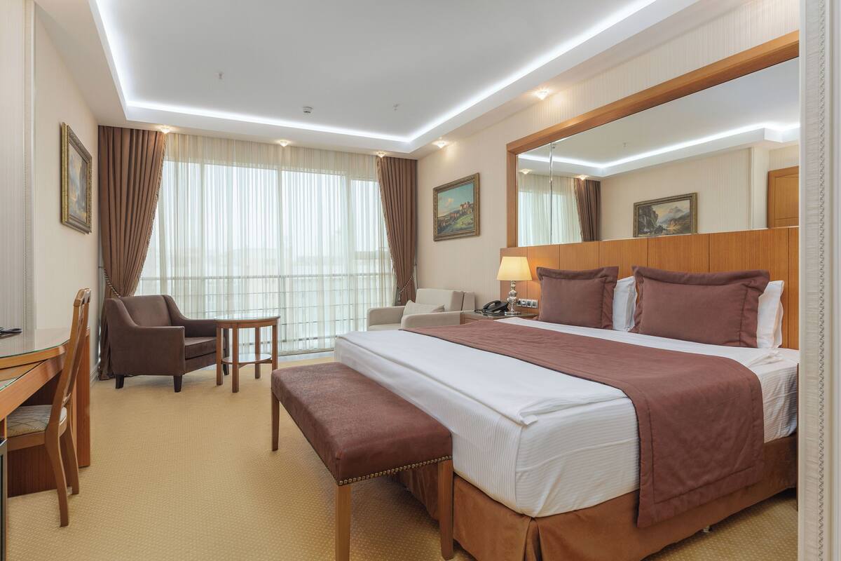 A hotel room with a large bed, a desk with a chair, a small table, and a bench.
